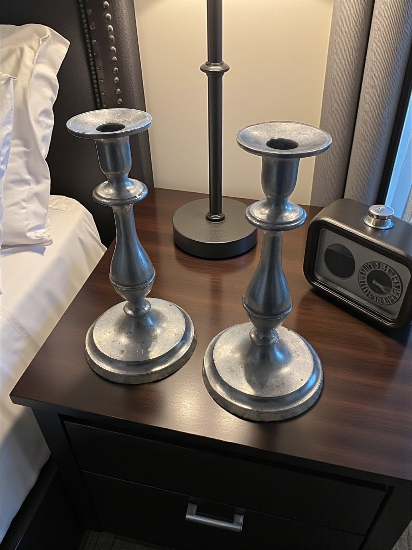Two silver candle holders on a wooden nightstand with a lamp and clock in the background.