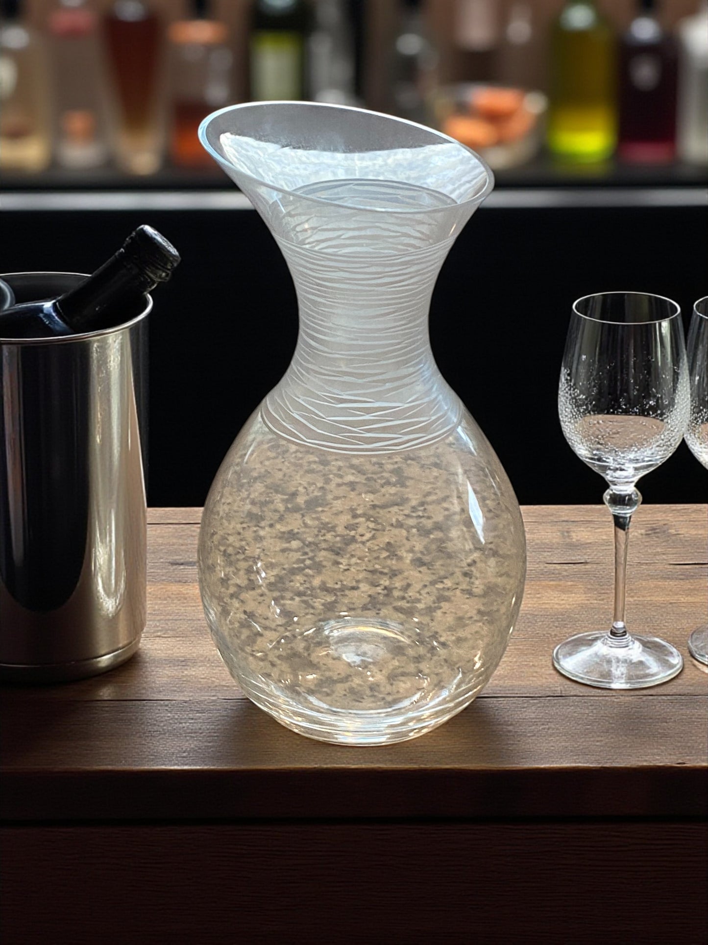 Clear glass decanter on a bar counter with wine glasses and a wine cooler in the background.
