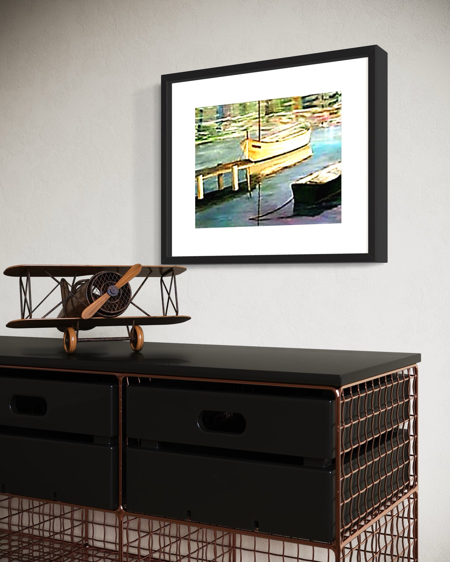 Framed artwork of a dock scene with boats on a wall above a black cabinet with a model airplane.