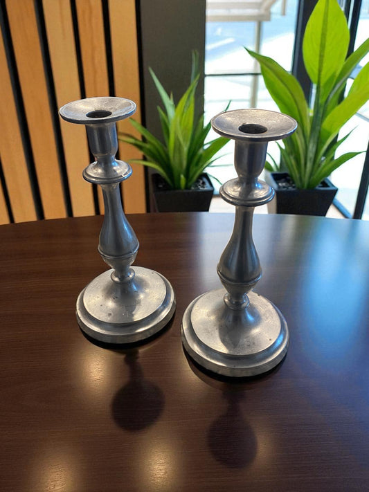 Two silver candle holders on a wooden surface with plants in the background.